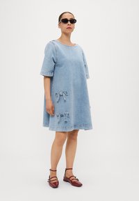 Vestido de ganga em azul claro, mangas curtas, silhueta oversized, com dois apliques de laço em ganga. Modelo usa óculos de sol pretos e sapatos vermelhos.