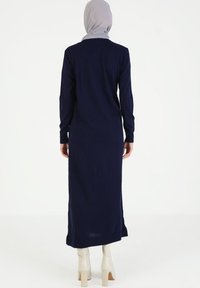 Abito lungo blu navy con maniche lunghe, texture a coste e spacco laterale, abbinato a stivali alla caviglia crema e una sciarpa grigia chiara.