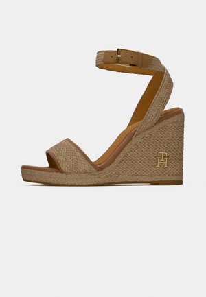 Sandale compensée en tan avec texture tressée, bride de cheville avec boucle dorée, et orteils ouverts, arborant un logo "TH" doré sur le talon.