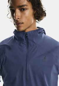 Giacca leggera navy con colletto alto e chiusura zip. Presenta un logo minimalista sul petto e un tessuto liscio e resistente all'acqua.