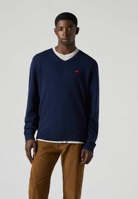 Marinblå v-ringad tröja i ett mjukt tyg, utrustad med en röd logotypbricka på bröstet, bärs över en ljusgrå skjorta och bruna byxor.