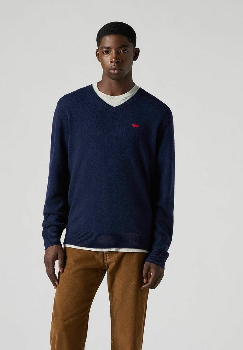 Marineblaues V-Ausschnitt-Sweatshirt aus weichem Material mit einem roten Logo-Patch auf der Brust, getragen über einem hellgrauen Hemd und braunen Hosen.