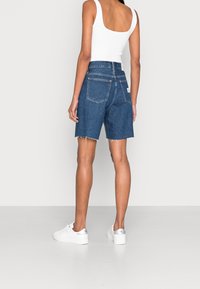 Jeansshorts i mörkblått med slitna kanter, två bakfickor, i kombination med en vit linne och vita sneakers med silverdetaljer.