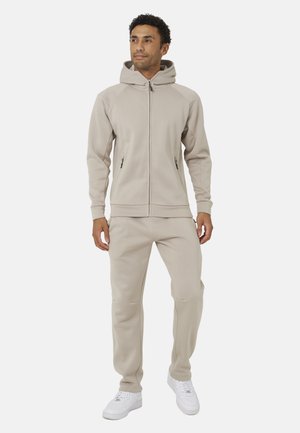Hombre de pie con una sudadera beige con cremallera y pantalones deportivos a juego, con zapatillas blancas sobre un fondo claro y liso.