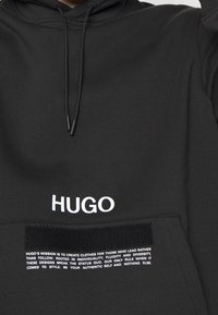 Zwarte fleece hoodie met een trekkoord in de capuchon en een groot wit "HUGO" logo. Bevat een gestructureerde zwarte strook met bedrukte witte tekst.
