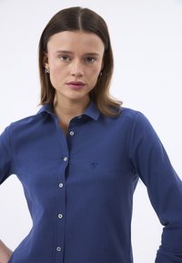 Blaues Hemd aus glattem Stoff mit Knopfleiste, das auf der linken Brust ein kleines gesticktes Vogel-Logo und silberne Knöpfe aufweist.