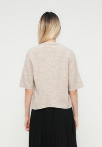 Beige kurzärmeliger Pullover mit weicher Textur, rundem Ausschnitt und lockerer Passform. Mit einem schwarzen Faltenrock getragen, von hinten betrachtet.