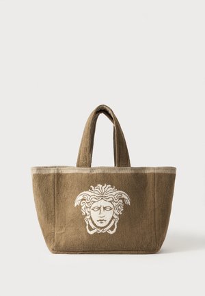 Brown tote bag with thick handles featuring a white Medusa head design on the front, set against a plain light background.
