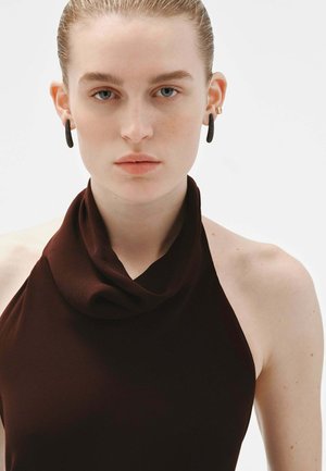 Top halter con cuello tipo cowl en color marrón con una textura suave. El modelo lleva pendientes de aro pequeños. El fondo neutro resalta la simplicidad de la prenda.