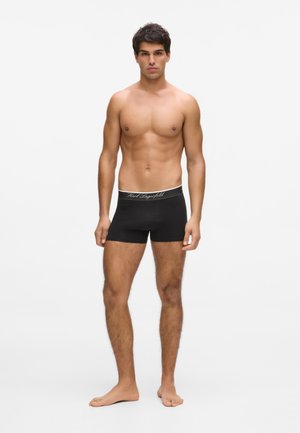 Homme aux cheveux courts et foncés, debout pieds nus, portant un boxer noir avec le logo "Karl Lagerfeld" sur la ceinture, sur un fond blanc.