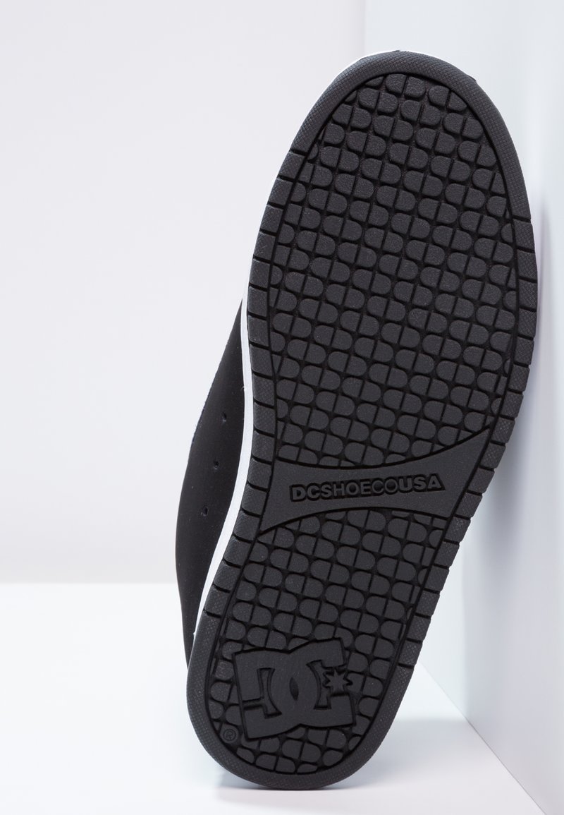 Suela de zapatilla de goma negra con patrón de agarre texturizado, que presenta una entresuela blanca. Detalle del logo grabado cerca del talón.