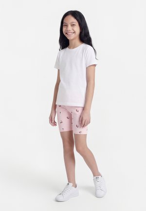 Fille souriante debout, portant un t-shirt blanc, un short rose à motifs cerises et des baskets blanches, sur fond blanc uni.