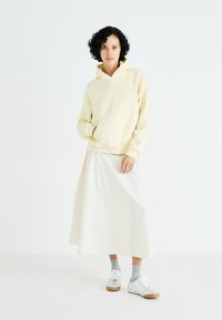 Light yellow hoodie with a front pocket, paired with a flowing white skirt, gray socks, and silver sneakers, set against a plain background.