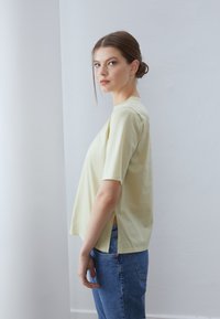 Tricou din bumbac de culoare galben deschis, cu mâneci scurte, fante laterale și o croială lejeră, asortat cu blugi albaștri. Design simplu și textură netedă.
