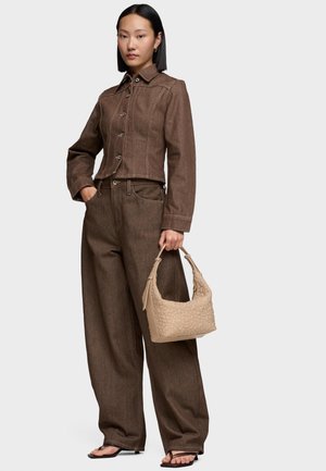Mujer con chaqueta de mezclilla marrón y pantalones de pierna ancha, sosteniendo un bolso beige con textura, de pie con una mano en el bolsillo y sandalias descalzas.