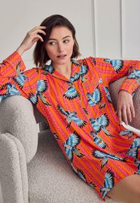 Woman reclining on textured armchair wearing bright orange button-up shirt dress patterned with blue parrots and pink diagonal stripes.