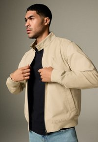 Veste beige zippée avec un col montant et des poignets côtelés, portée sur un t-shirt sombre. Présente un logo discret sur la poitrine et des poches latérales.