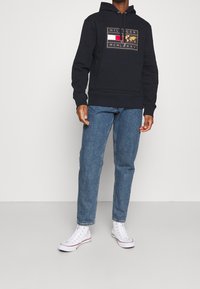 Námořnická mikina s kapucí s grafickým motivem na přední straně, obsahující červené a bílé prvky. Kombinováno s světle modrými džínami a bílými teniskami.