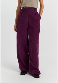 Wijde corduroy broek in diep paars, met een gestructureerde afwerking, hoge taille met plooien en zijzakken. Gecombineerd met donkere schoenen.