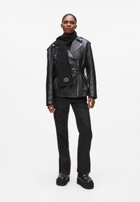 Veste de motard en cuir noir avec ceinture, texture côtelée et accents brodés. Associée à une écharpe noire, un pantalon noir ajusté et des bottes à plateforme.