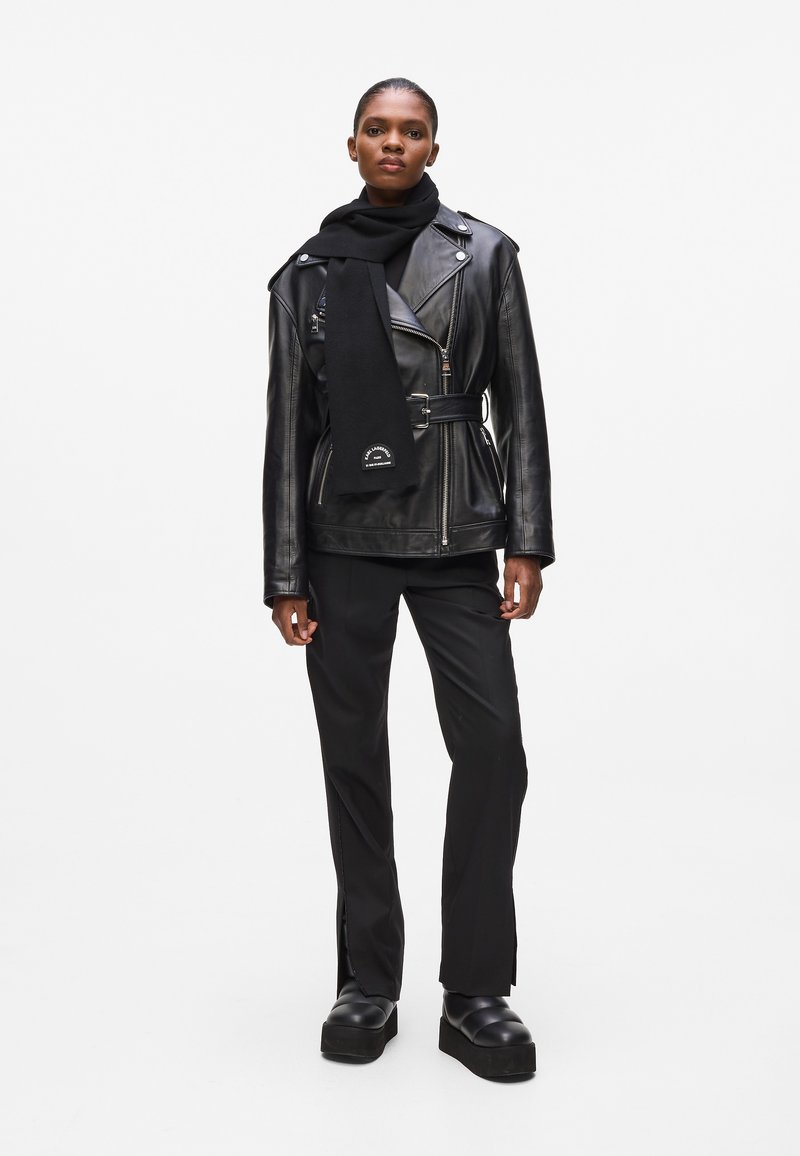 Veste de motard en cuir noir avec ceinture, texture côtelée et accents brodés. Associée à une écharpe noire, un pantalon noir ajusté et des bottes à plateforme.