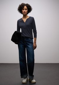 Vrouw met kort krullend haar, gekleed in een donker henley-shirt met lange mouwen, wijd omgeslagen broekspijpen, witte sneakers, en een zwarte jas vasthoudend.