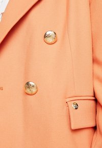 Blazer v broskvové barvě s texturovaným povrchem, se dvěma velkými zlatými knoflíky a malým akcentovým knoflíkem s logem poblíž kapsy.