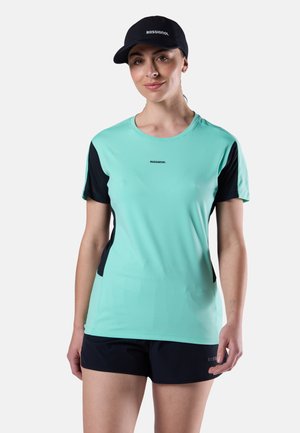 Woman wearing a black Rossignol cap, mint green and black Rossignol short-sleeve shirt, and black Rossignol shorts, standing against white background.