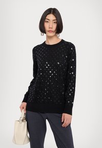 Schwarzer Pullover mit einem strukturierten Design aus kreisförmigen Pailletten, gerippten Bündchen und Rundhalsausschnitt. Kombiniert mit einer hellen Handtasche.