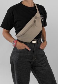 Borsa a tracolla beige realizzata in materiale morbido, con chiusura a zip, tracolla regolabile e un branding discreto sul davanti.