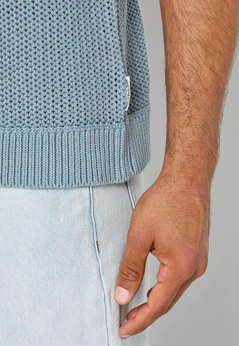 Helles blaues gestricktes Sweatshirt mit einem strukturierten Muster, geripptem Saum und einem kleinen Etikett an der Seite. Kombiniert mit hellen Jeans.