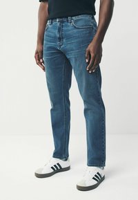 Jean en denim bleu foncé avec une coupe droite, doté de cinq poches et d'un zip, associé à des baskets blanches avec des rayures noires.