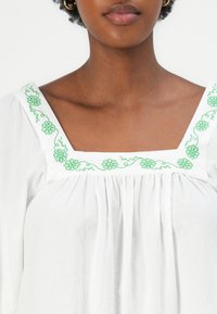 Witte blouse met een vierkante halslijn, versierd met groene bloemenborduursels langs de kraag. De textuur lijkt zacht en lichtgewicht.