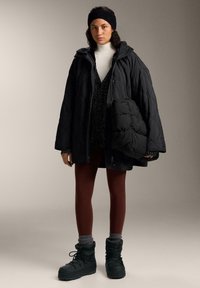 Black quilted outer jacket with hood, layered over a dark sweater, paired with burgundy leggings and green winter boots. Includes a puffy handbag.