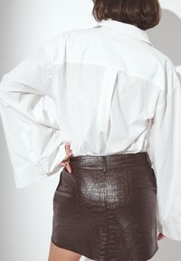 Valge pikkade varrukatega särk, millel on lõtv lõige, millele on sobitatud ülikitsas pruun krokodillimustriga nahkseelik, millel on keskmine õmblus.