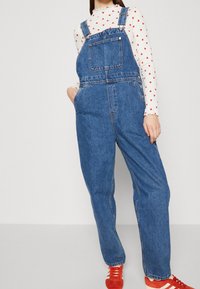 Salopette in denim di un blu medio con spalline regolabili, tasca frontale e gambe larghe. Abbinata a una camicia bianca a maniche lunghe con cuori rossi.