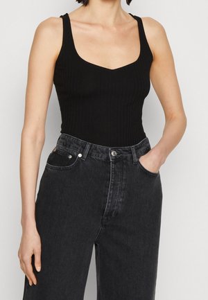 Kobieta ubrana w czarny prążkowany, bezrękawnik wsunięty w wysokowzrostą czarną dżinsową spódnicę, z jedną ręką w kieszeni, stojąca na tle jednolitego tła.