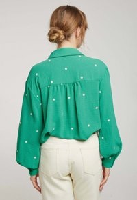 Blouse verte en tissu texturé, avec des détails de broderie florale et une coupe ample. Équipée d'un col et de poignets boutonnés.