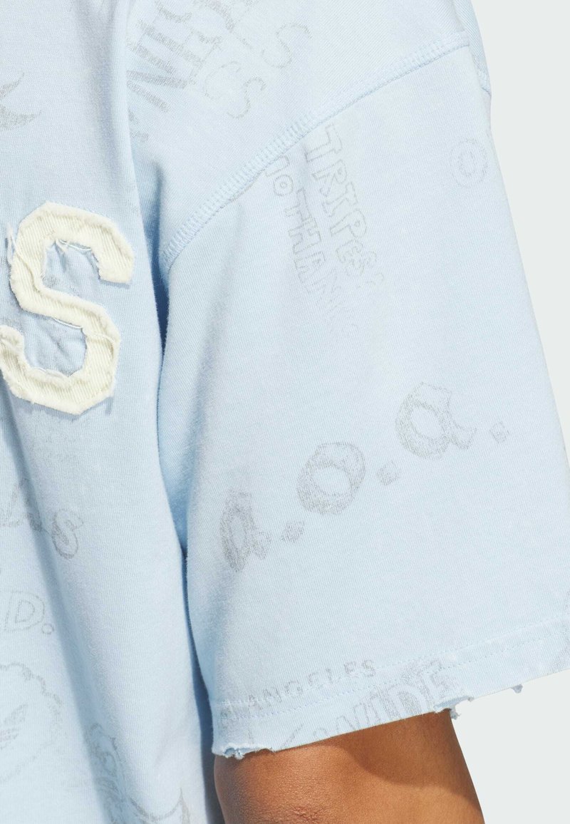 Camisa azul claro de manga corta con patrones de texto gris desvaído y una aplicación blanca con textura de la letra "S" cerca del hombro.