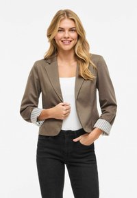 Femme souriante aux longs cheveux blonds portant un blazer marron court sur un haut blanc, un jean noir, posant avec une main dans la poche.