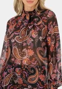 Luźna czarna bluzka z wzorem paisley w pomarańczowym i fioletowym, z zawiązanym dekoltem i długimi, drapowanymi rękawami. Lekki, prześwitujący materiał.