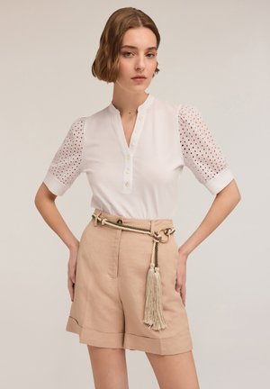 Blusa bianca con colletto e chiusura a bottoni, con dettagli a occhio di pernice sulle maniche a sbuffo. Abbinata a pantaloni corti beige a vita alta e una cintura con nappe.