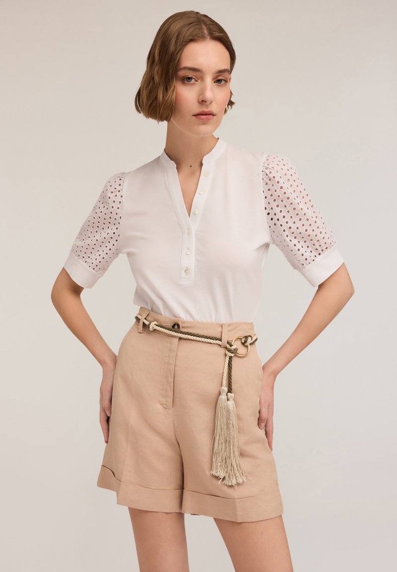 Witte blouse met een kraag en knoopopening, met oogjesdetail op de pofmouwen. Gecombineerd met beige hooggesneden shorts en een kwastgordel.