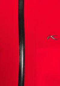 Giacca rossa con cerniera nera, tessuto liscio e logo metallico in evidenza sul lato. La cerniera è verticale.