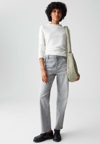 Frau mit einem weißen strukturierten Langarmoberteil und hellgrauen Jeans, die eine gesteppte olivgrüne Schultertasche trägt, steht vor einem neutralen Hintergrund.