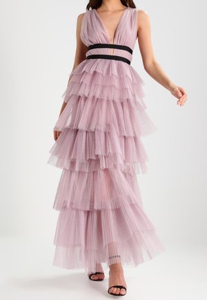 Kobieta w bezrękawowej, warstwowej sukni z jasnofioletowego tiulu z czarnymi pasami w talii i czarnych sandałach na odkryte palce, idąca na tle białego tła.