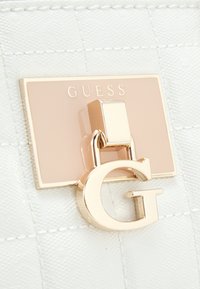 Witte getextureerde materiaal met een goudkleurige hardware-detail. Heeft een rechthoekige plaquette met "GUESS" en een opvallend gouden "G" accent.