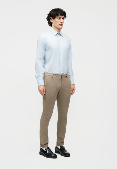 Νέος άνδρας που στέκεται, φοράει ανοιχτό μπλε πουκάμισο με ρίγες, μπεζ παντελόνι στενής γραμμής, λευκές κάλτσες και μαύρα δερμάτινα μοκασίνια, μπροστά από απλό φόντο.