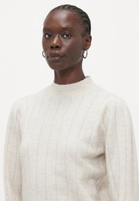 Maglione color beige chiaro con collo alto, caratterizzato da una texture a righe verticali e dettagli di cucitura discreti. Il modello ha i capelli intrecciati e indossa orecchini a cerchio.
