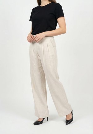 Donna con top nero a maniche corte, pantaloni beige a gamba larga e vita alta, e tacchi neri con punta appuntita, in piedi davanti a uno sfondo bianco.